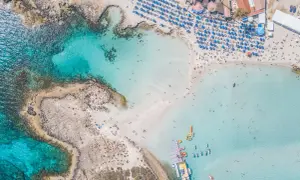 Piękna plaża z turkusową wodą, leżakami i parasolami. Widać łodzie i ludzi kąpiących się w morzu. To idealne miejsce na wakacje, oferujące wiele atrakcji w Ayia Napa.