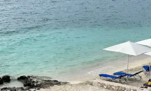 Piękna pogoda w Sarandzie: turkusowe morze, piaszczysta plaża z leżakami i parasolami, pomarańczowe boje na wodzie.