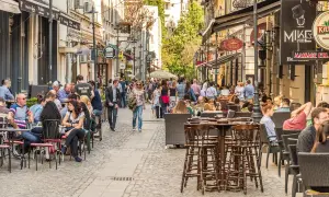 Tętniąca życiem ulica w Bukareszcie, pełna ludzi siedzących w kawiarniach i restauracjach. W tle widać zabytkowe budynki.