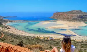 Widok na turkusowe wody i piaszczyste **kreta plaże** z punktu widokowego. Kobieta siedzi na skale, podziwiając krajobraz.