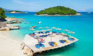 Słoneczny dzień na Ksamil Beach. Drewniany pomost z leżakami i parasolami, turkusowa woda i zielona wyspa w tle.