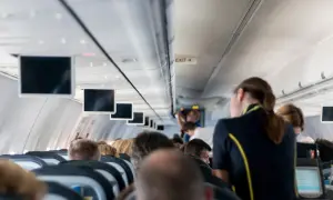 Wnętrze samolotu, rzędy siedzeń, ekrany nad głowami. Klaustrofobia a lot samolotem? Niektórzy pasażerowie wyglądają na zaniepokojonych.