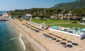 Słoneczna plaża Baia Salima Kemer z leżakami i parasolami, zielony trawnik hotelowy i góry w tle.