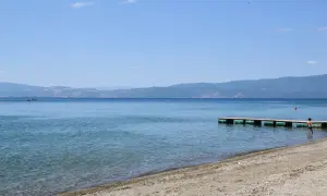 Spokojne jezioro Ochrydzkie z piaszczystą plażą i drewnianym pomostem. W oddali góry, na wodzie widać kajak.
