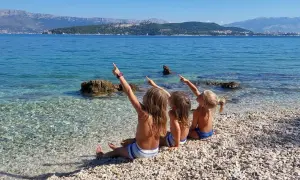 Trzy dziewczynki siedzą na kamienistej plaży w Chorwacji, wskazując palcami na morze i góry. Idealne miejsce, gdzie do Chorwacji z dziećmi.