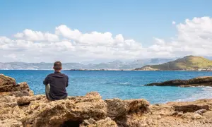 Mężczyzna siedzi na skale, patrząc na morze. Kreta pogoda marzec, błękitne niebo z chmurami, spokojne morze.