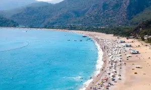 Tłum ludzi wypoczywa na piaszczystej plaży nad turkusowym morzem egejskim, otoczonej zielonymi wzgórzami.