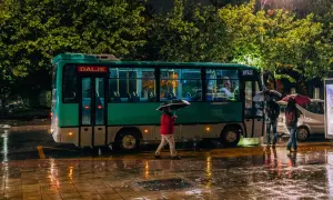 Deszczowa noc w Tiranie. Autobus z napisem "Dalje" i "Hyrje" stoi na mokrej ulicy, ludzie z parasolami czekają na przystanku.
