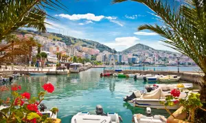 Albania, lipiec: słoneczna pogoda nad zatoką z łodziami i palmami. Miasto na wzgórzu w tle.