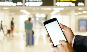 Ręka trzyma smartfon z pustym ekranem, gotowa do okazania lotniczego biletu elektronicznego. W tle lotnisko z tablicami odlotów i ludźmi.