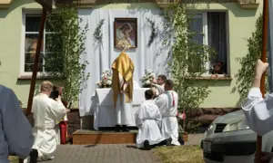 Uroczystość Bożego Ciała w długi weekend czerwiec. Procesja przed domem, kapłan w złotej szacie, wierni klęczą.