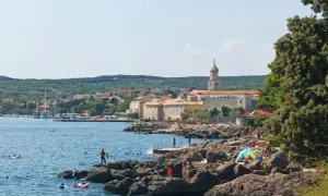 Słoneczny dzień na wyspie Krk. Ludzie odpoczywają na skalistej plaży, kąpią się w morzu. W tle widać miasteczko z charakterystyczną wieżą.