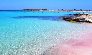 Rajska plaża na Krecie z różowym piaskiem i krystalicznie czystą, turkusową wodą. W tle widać ludzi kąpiących się i odpoczywających.