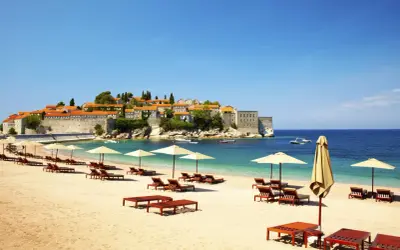Idealne czarnogóra wakacje all inclusive: piaszczysta plaża, leżaki, parasole i widok na urokliwe miasteczko na wyspie.