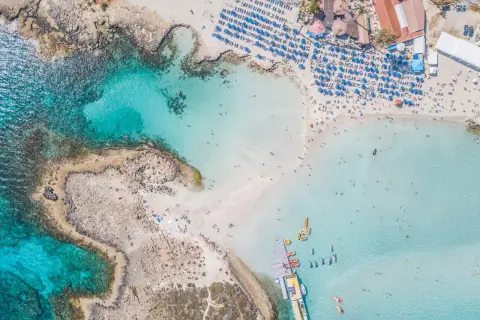 Piękna plaża z turkusową wodą, leżakami i parasolami. Widać łodzie i ludzi kąpiących się w morzu. To idealne miejsce na wakacje, oferujące wiele atrakcji w Ayia Napa.