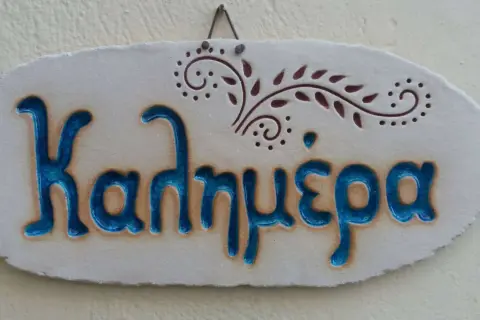 Ceramiczna tabliczka z napisem "Καλημέρα" (do zobaczenia po grecku) i ozdobnym motywem roślinnym.