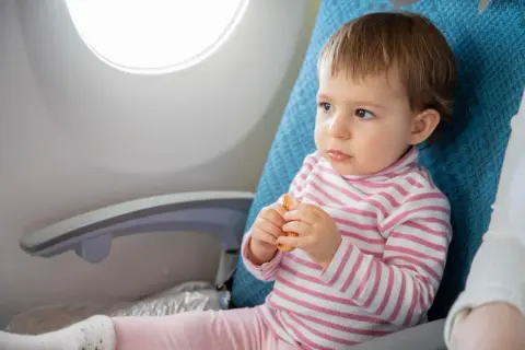Małe dziecko z zaciekawieniem patrzy przez okno samolotu, trzymając w rączkach ciasteczko. To jego pierwszy lot samolotem z dzieckiem 3 letnim.