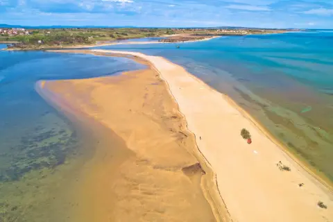 Chorwacja plaże piaszczyste, piaszczysta mierzeja otoczona turkusową wodą, w oddali widać zabudowania.