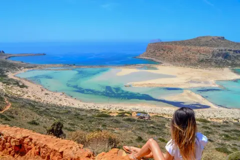 Widok na turkusowe wody i piaszczyste **kreta plaże** z punktu widokowego. Kobieta siedzi na skale, podziwiając krajobraz.