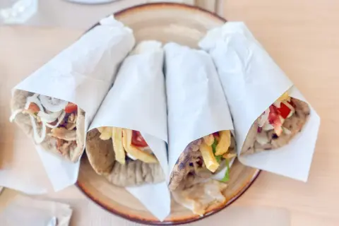 Cztery pyszne souvlaki gyros z mięsem, frytkami, warzywami i sosem, zawinięte w papier, na talerzu.
