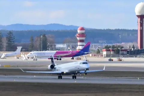 Samolot kołuje po płycie lotniska, gotowy do pierwszego lotu samolotem. W tle różowy samolot Wizz Air i wieża kontroli lotów.