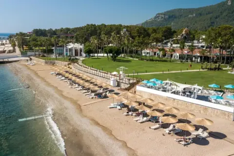 Słoneczna plaża Baia Salima Kemer z leżakami i parasolami, zielony trawnik hotelowy i góry w tle.