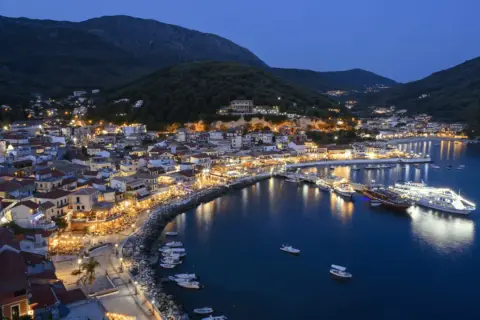 Widok na oświetlone miasto Parga nad morzem o zmierzchu, z górami w tle.