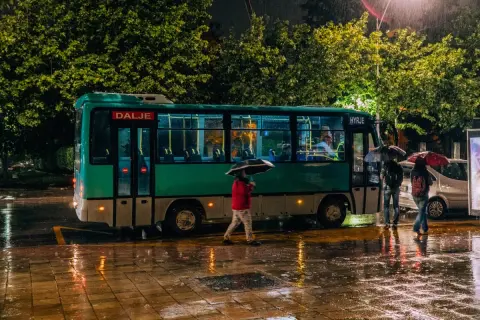 Deszczowa noc w Tiranie. Autobus z napisem "Dalje" i "Hyrje" stoi na mokrej ulicy, ludzie z parasolami czekają na przystanku.