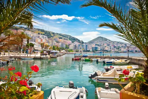 Albania, lipiec: słoneczna pogoda nad zatoką z łodziami i palmami. Miasto na wzgórzu w tle.
