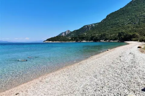 Chorwacja piaszczyste plaże blisko Polski. Dwie osoby pływają w krystalicznie czystej wodzie przy kamienistej plaży, w tle zielone wzgórza.