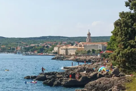 Słoneczny dzień na wyspie Krk. Ludzie odpoczywają na skalistej plaży, kąpią się w morzu. W tle widać miasteczko z charakterystyczną wieżą.