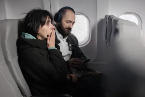 Kobieta z nerwicą lękową w samolocie zakrywa usta dłonią, próbując opanować strach. Obok siedzi mężczyzna w słuchawkach.