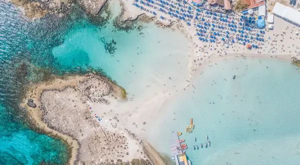 Piękna plaża z turkusową wodą, leżakami i parasolami. Widać łodzie i ludzi kąpiących się w morzu. To idealne miejsce na wakacje, oferujące wiele atrakcji w Ayia Napa.