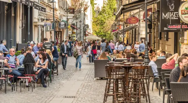 Tętniąca życiem ulica w Bukareszcie, pełna ludzi siedzących w kawiarniach i restauracjach. W tle widać zabytkowe budynki.