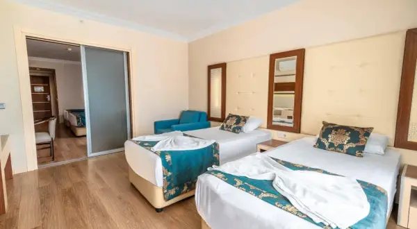 Pokój hotelowy z dwoma łóżkami, ozdobiony turkusowymi narzutami. Widok na sąsiedni pokój przez przesuwne drzwi. Idealne miejsce na relaks po dniu spędzonym w beach club doganay.