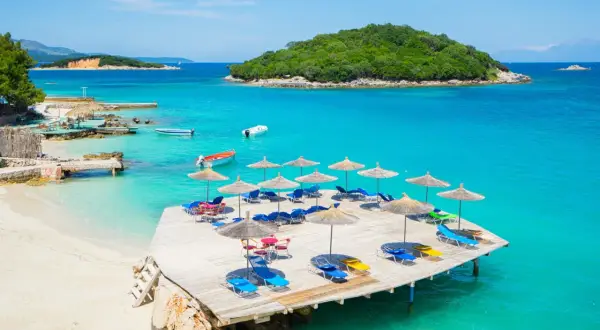 Słoneczny dzień na Ksamil Beach. Drewniany pomost z leżakami i parasolami, turkusowa woda i zielona wyspa w tle.
