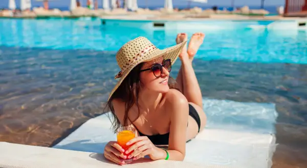 Kobieta w słomkowym kapeluszu i okularach przeciwsłonecznych relaksuje się przy basenie z drinkiem. Idealny urlop all inclusive.