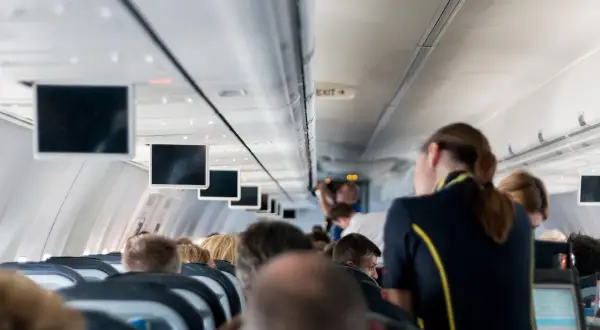 Wnętrze samolotu, rzędy siedzeń, ekrany nad głowami. Klaustrofobia a lot samolotem? Niektórzy pasażerowie wyglądają na zaniepokojonych.
