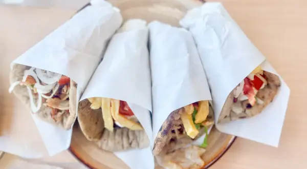 Cztery pyszne souvlaki gyros z mięsem, frytkami, warzywami i sosem, zawinięte w papier, na talerzu.