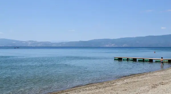Spokojne jezioro Ochrydzkie z piaszczystą plażą i drewnianym pomostem. W oddali góry, na wodzie widać kajak.