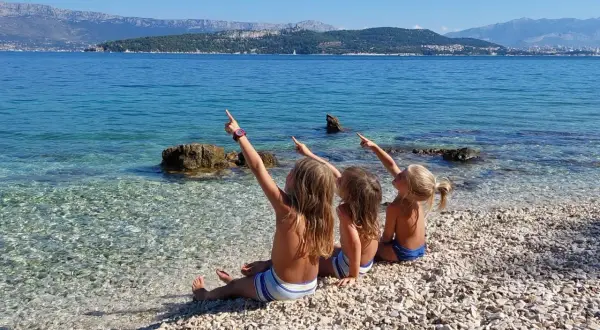 Trzy dziewczynki siedzą na kamienistej plaży w Chorwacji, wskazując palcami na morze i góry. Idealne miejsce, gdzie do Chorwacji z dziećmi.