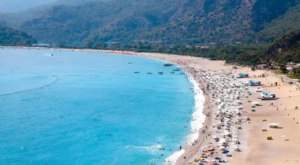 Tłum ludzi wypoczywa na piaszczystej plaży nad turkusowym morzem egejskim, otoczonej zielonymi wzgórzami.