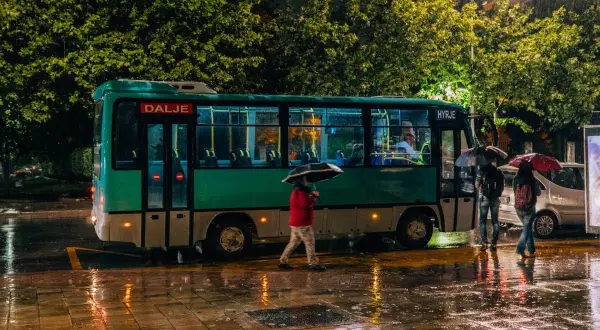 Deszczowa noc w Tiranie. Autobus z napisem "Dalje" i "Hyrje" stoi na mokrej ulicy, ludzie z parasolami czekają na przystanku.