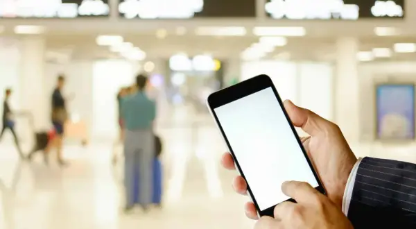 Ręka trzyma smartfon z pustym ekranem, gotowa do okazania lotniczego biletu elektronicznego. W tle lotnisko z tablicami odlotów i ludźmi.