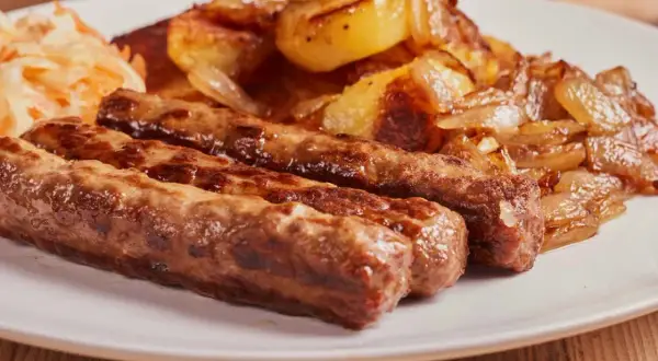 Trzy soczyste ćevapi, klasyka kuchni czarnogórskiej, podane z pieczonymi ziemniakami i cebulą oraz surówką.