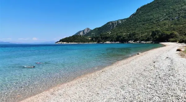 Chorwacja piaszczyste plaże blisko Polski. Dwie osoby pływają w krystalicznie czystej wodzie przy kamienistej plaży, w tle zielone wzgórza.
