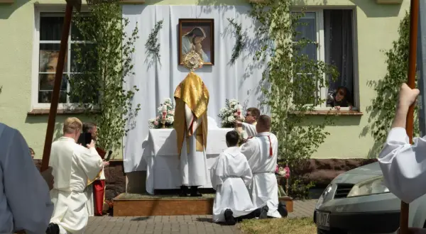 Uroczystość Bożego Ciała w długi weekend czerwiec. Procesja przed domem, kapłan w złotej szacie, wierni klęczą.