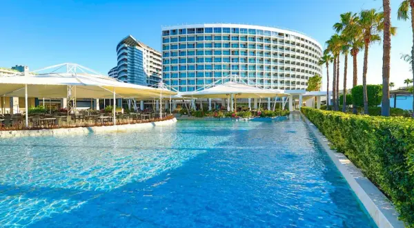 Luksusowy hotel z basenem i palmami. W oddali widać budynek w kształcie fali, a bliżej restaurację pod białymi markizami. Idealne miejsce na wypoczynek w Crystal Centro.