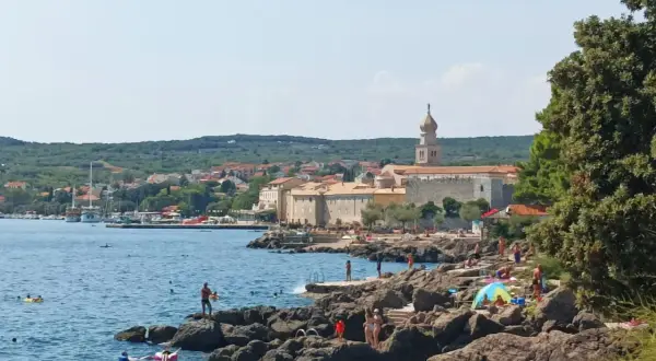 Słoneczny dzień na wyspie Krk. Ludzie odpoczywają na skalistej plaży, kąpią się w morzu. W tle widać miasteczko z charakterystyczną wieżą.