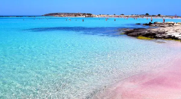 Rajska plaża na Krecie z różowym piaskiem i krystalicznie czystą, turkusową wodą. W tle widać ludzi kąpiących się i odpoczywających.